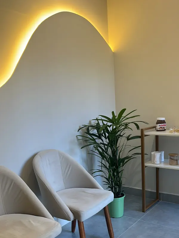 Recepção com duas poltronas creme, uma planta e a parede recortada em onda com luz amarela por trás